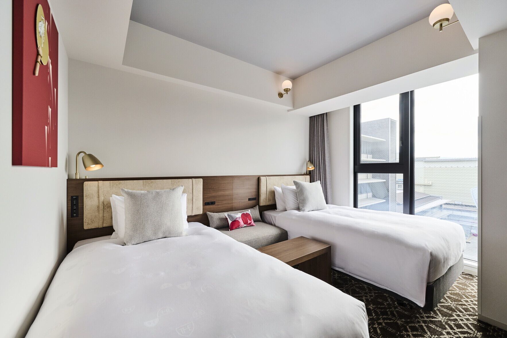 A hotel room with two beds, a wooden headboard, a small table, a red artwork, and a large window offering a view.
