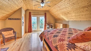 Una camera da letto mansardata accogliente, con soffitto in legno, un grande letto con copriletto, una sedia in vimini e vista all'esterno attraverso una porta in vetro.