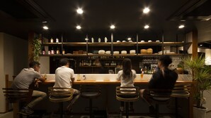 Zona de bar con taburetes de madera, estanterías con botellas y vasos, y una planta en maceta.