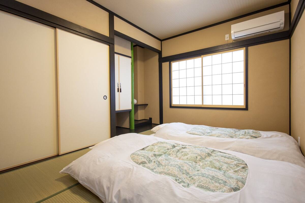 A traditional Japanese room with tatami flooring, two beds with patterned bedding, a sliding door, a window with a grid pattern, and an air conditioning unit.