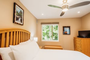 Ein Schlafzimmer mit Bett, Deckenventilator, Fenster mit Blick auf Grünflächen, Kommode und Fernseher.