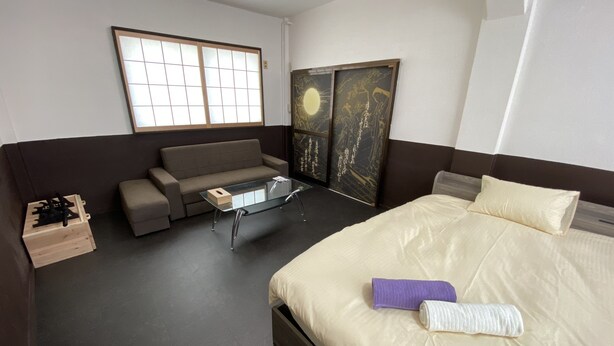 A hotel room with a bed, a sofa, a glass coffee table, a wooden chest, and a window with a view of the sky. - Yado KUKAI Shin Imamiya (Osaka)