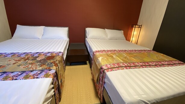 A hotel room with two single beds, each with a patterned bedspread, a small wooden nightstand, and a wall-mounted lamp.