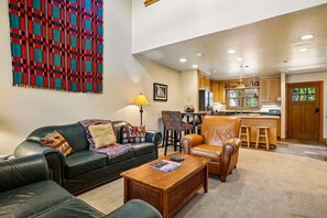A living room with a sofa, coffee table, and a patterned rug. There is a kitchen area with wooden cabinets and a bar stool.