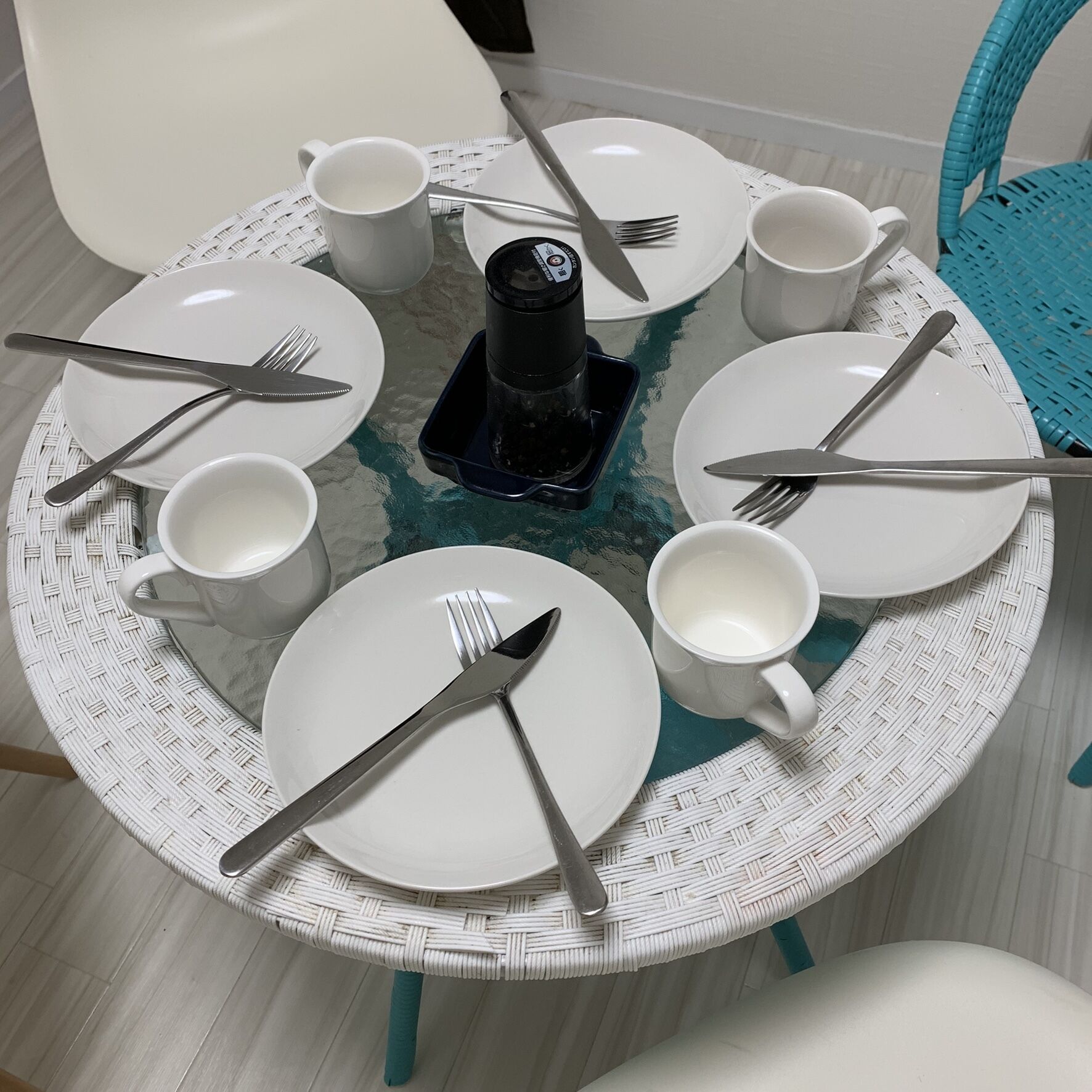 Una mesa redonda para comer con platos blancos, cubiertos y tazas blancas, centrada alrededor de una cafetera negra.