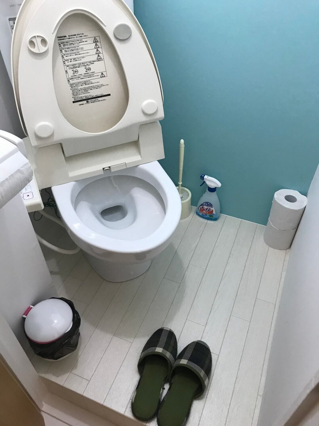 A bathroom with a toilet, a trash bin, a toilet brush, a spray bottle, and a pair of slippers on the floor.