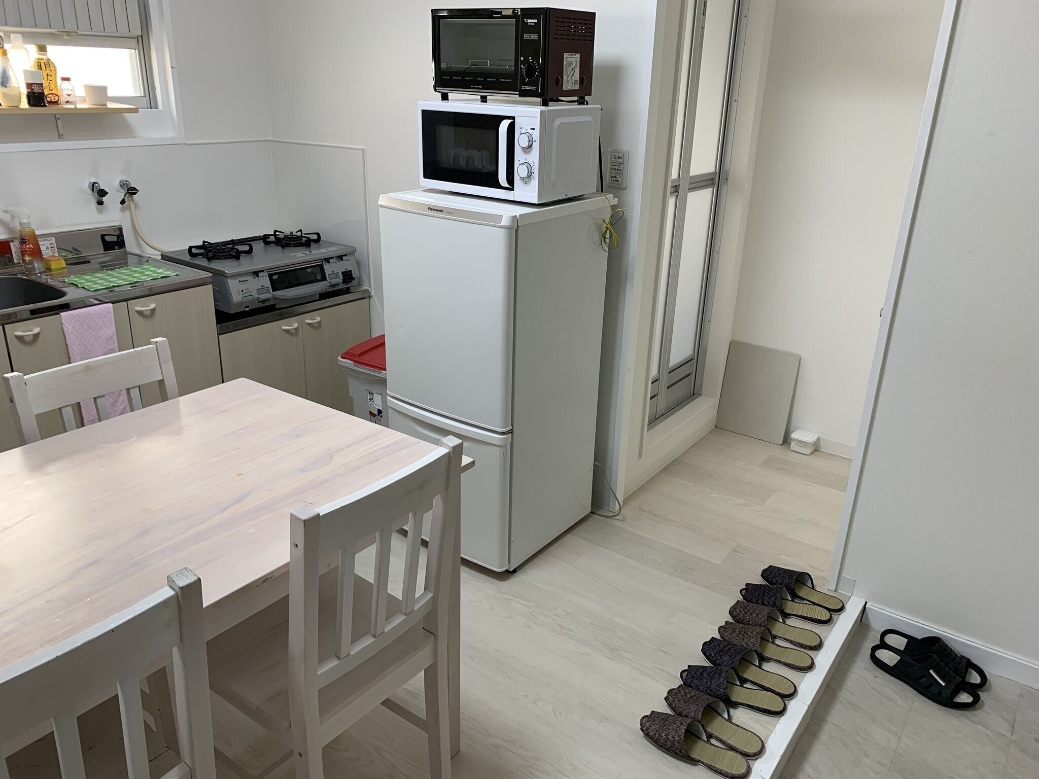 A compact kitchen with a refrigerator, microwave, and stove. There is a dining table with chairs, and a shelf with shoes and slippers.