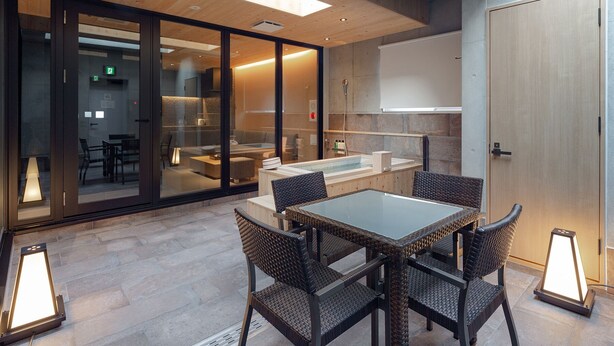 Un espacio interior moderno con separador de vidrio, techo de madera, zona de estar con sillas de mimbre y una pequeña piscina.