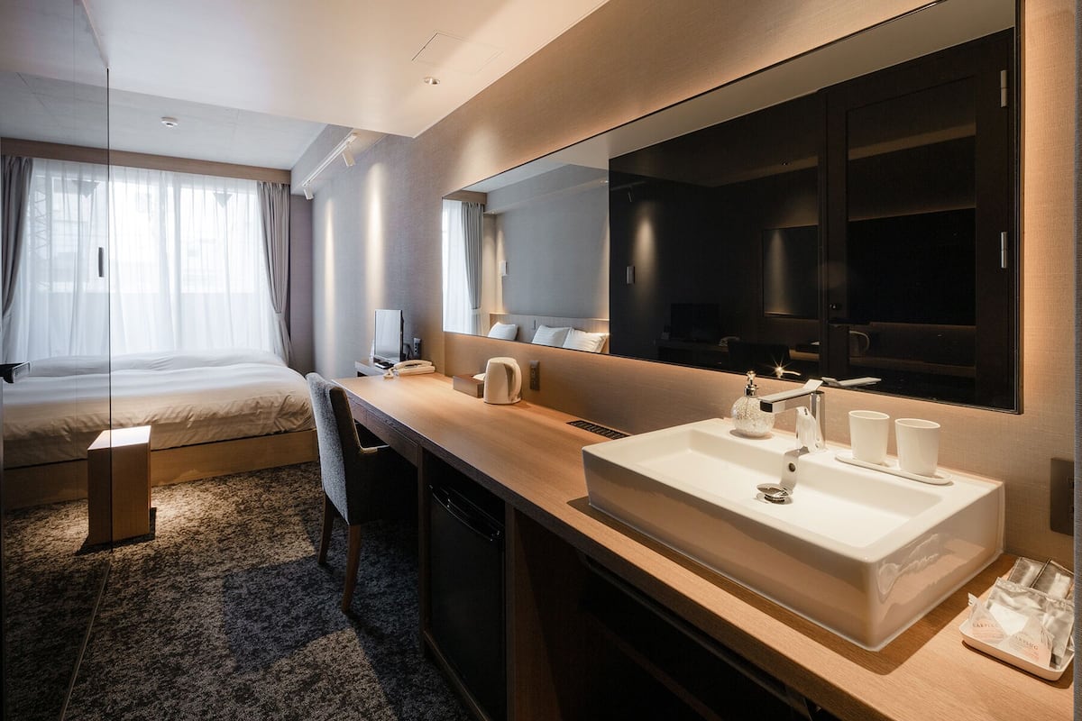 A modern hotel room with a large bed, a desk with a computer, a large mirror, and a bathroom with a sink and mirror.
