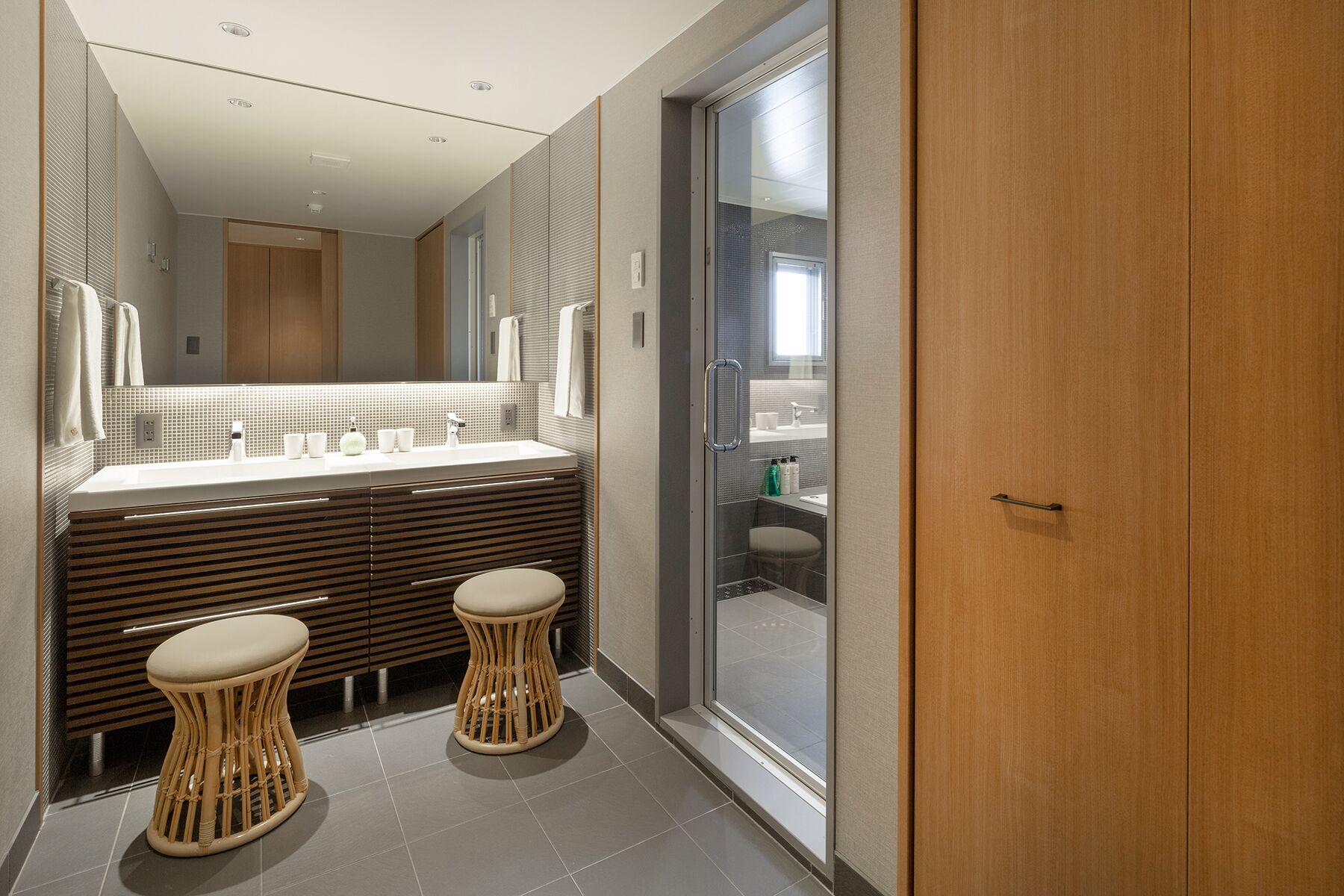A modern bathroom with a large mirror, wooden vanity, two stools, and a glass shower enclosure.