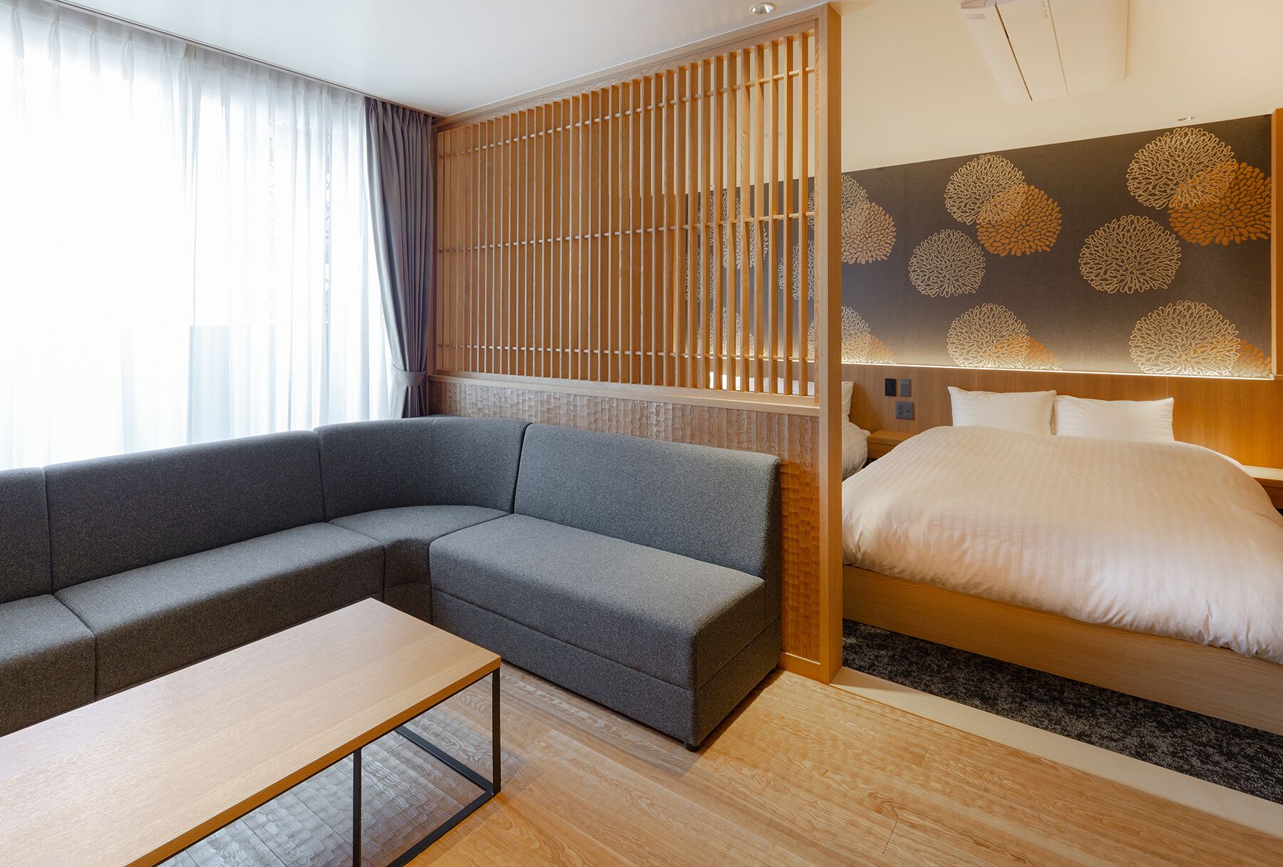 A modern hotel room with a grey sectional sofa, a wooden floor, a bed with white bedding, and a decorative wall with a floral pattern.
