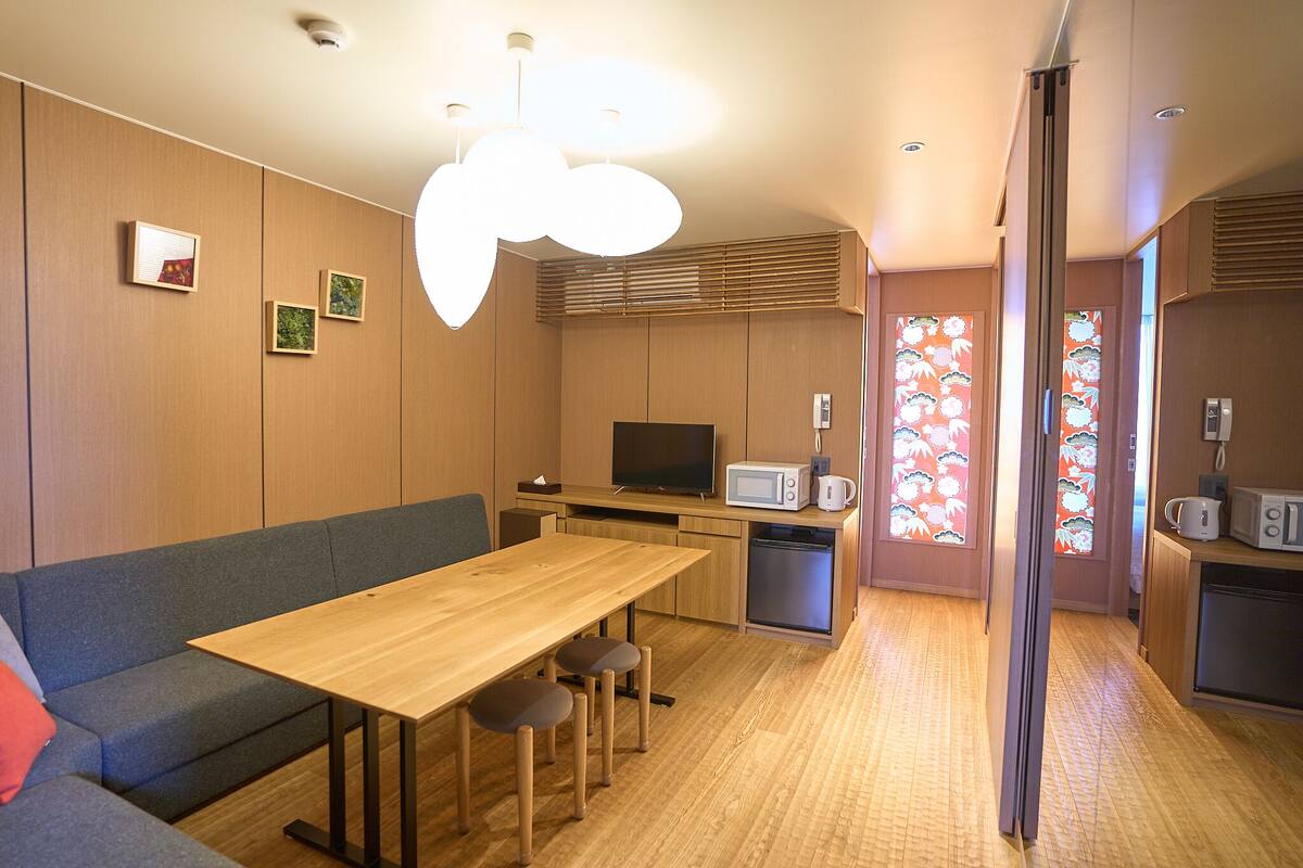 A compact living space with a wooden dining table, a sofa, a TV, and a kitchen area with a microwave and a refrigerator.