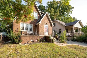 A two-story brick house with a front porch, a small garden, and a visible garage. - Ferndale Apartment w/ Yard - Near the Detroit Zoo! (Ferndale)