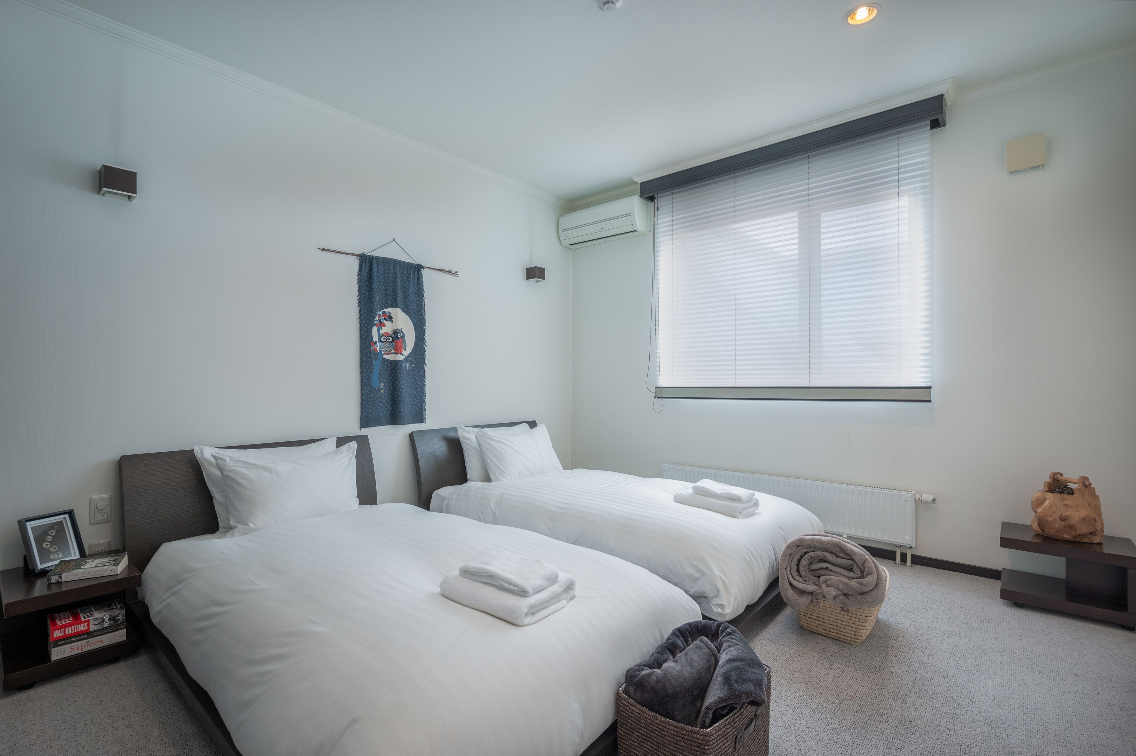Une chambre d’hôtel avec deux lits, un radiateur, une fenêtre avec des stores, une petite table et un climatiseur fixé au mur.