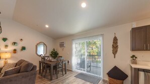 Una sala de estar moderna con sofá, mesa de comedor y sillas. Hay una zona de cocina con armarios y un refrigerador. Una puerta corrediza de vidrio da a un patio con vistas a la vegetación.