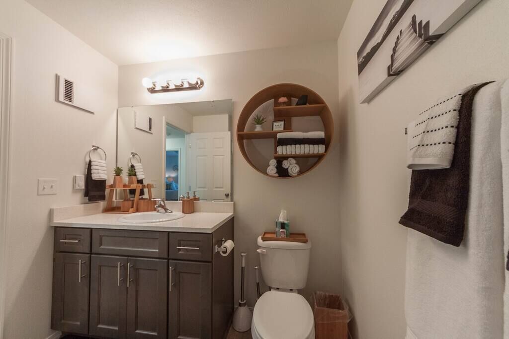 A bathroom with a toilet, sink, and vanity. There is a towel rack with towels and a shelf with toiletries.
