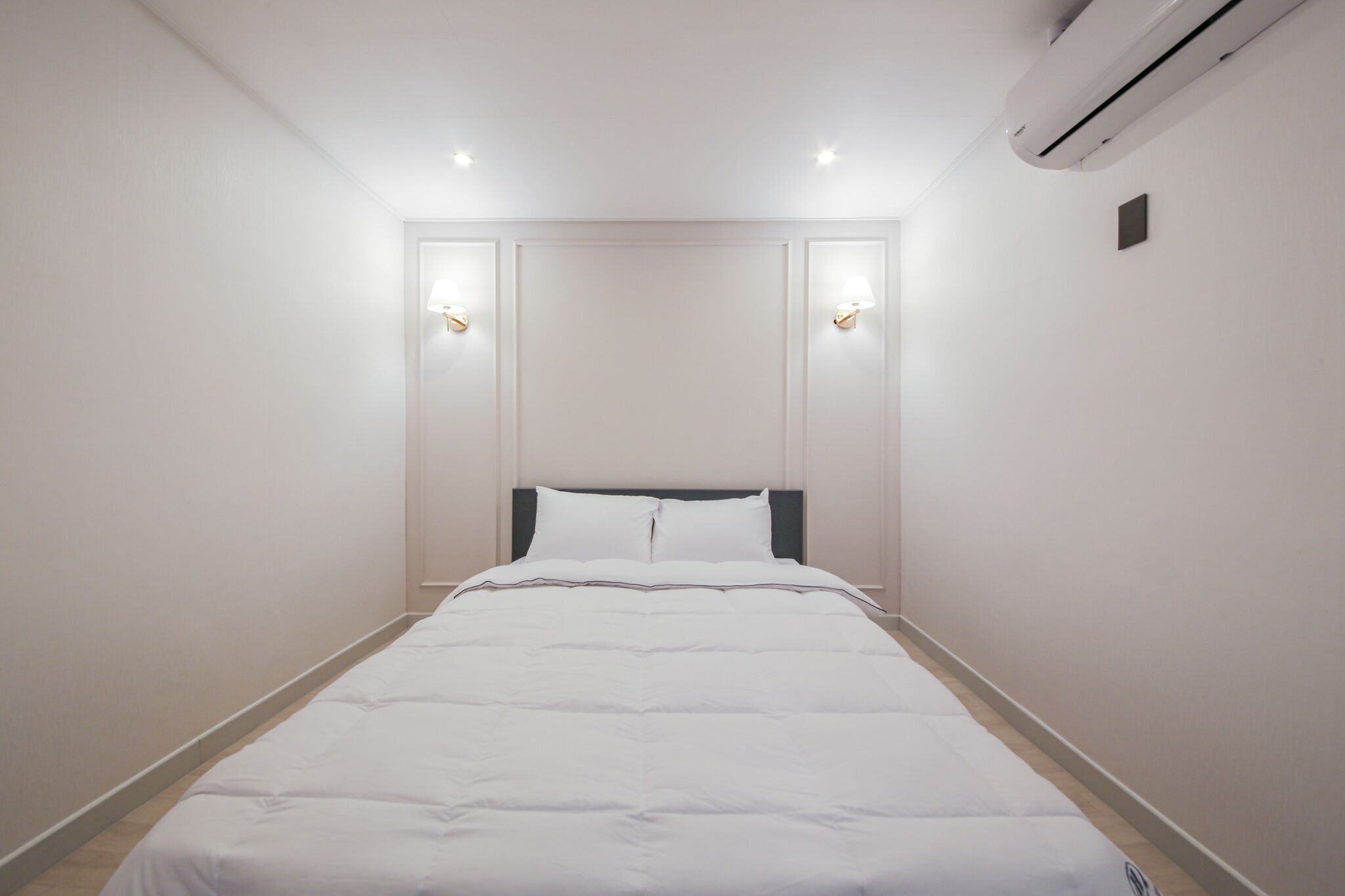 A neatly made bed in a hotel room with a headboard, two wall-mounted lights, and an air conditioning unit on the wall.