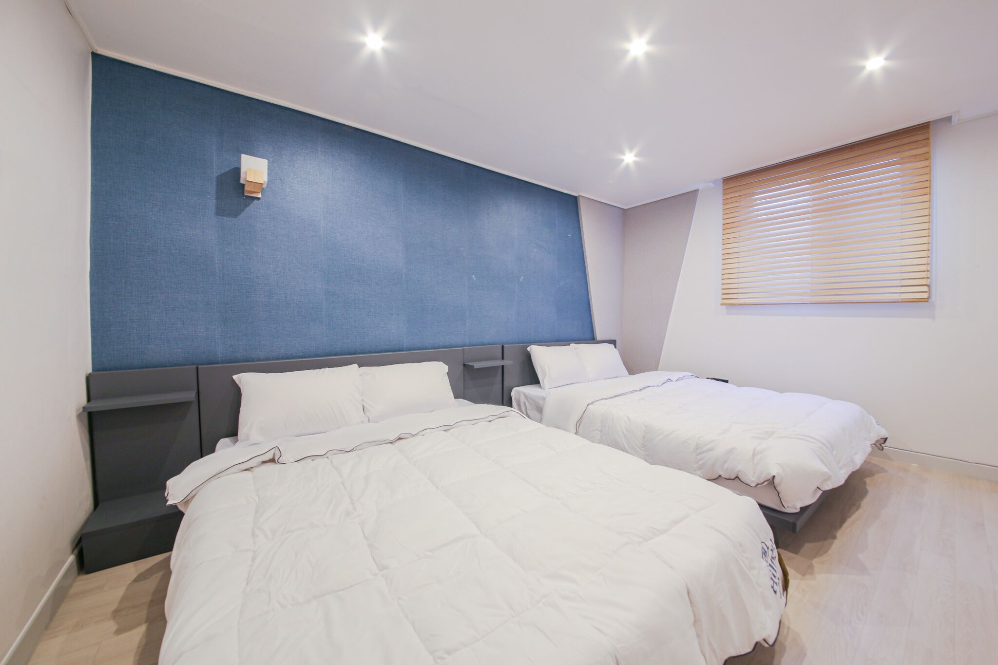 A modern hotel room with two beds, a blue accent wall, and a window with blinds.