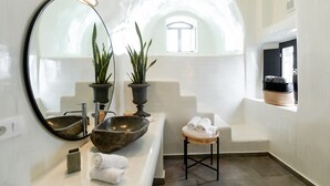 A modern bathroom with a large round mirror, a stone basin, two potted plants, a small stool with towels, and a window with a view of the sky. - Doukas Caldera Suites Fira (Santorini)