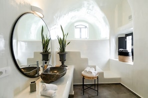 Experience the airy atmosphere of this bathroom with a vaulted ceiling, featuring a round mirror, stone basin sink, and lush greenery.