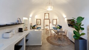 A modern kitchen with a white countertop, a dining area with a round table and chairs, and a large plant in a grey pot. - Doukas Caldera Suites Fira (Santorini)