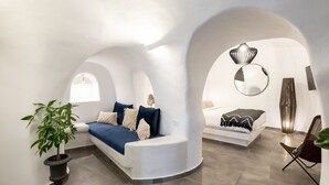 A modern interior with a curved white wall, a blue sofa, a bed, and a potted plant. - Doukas Caldera Suites Fira (Santorini)