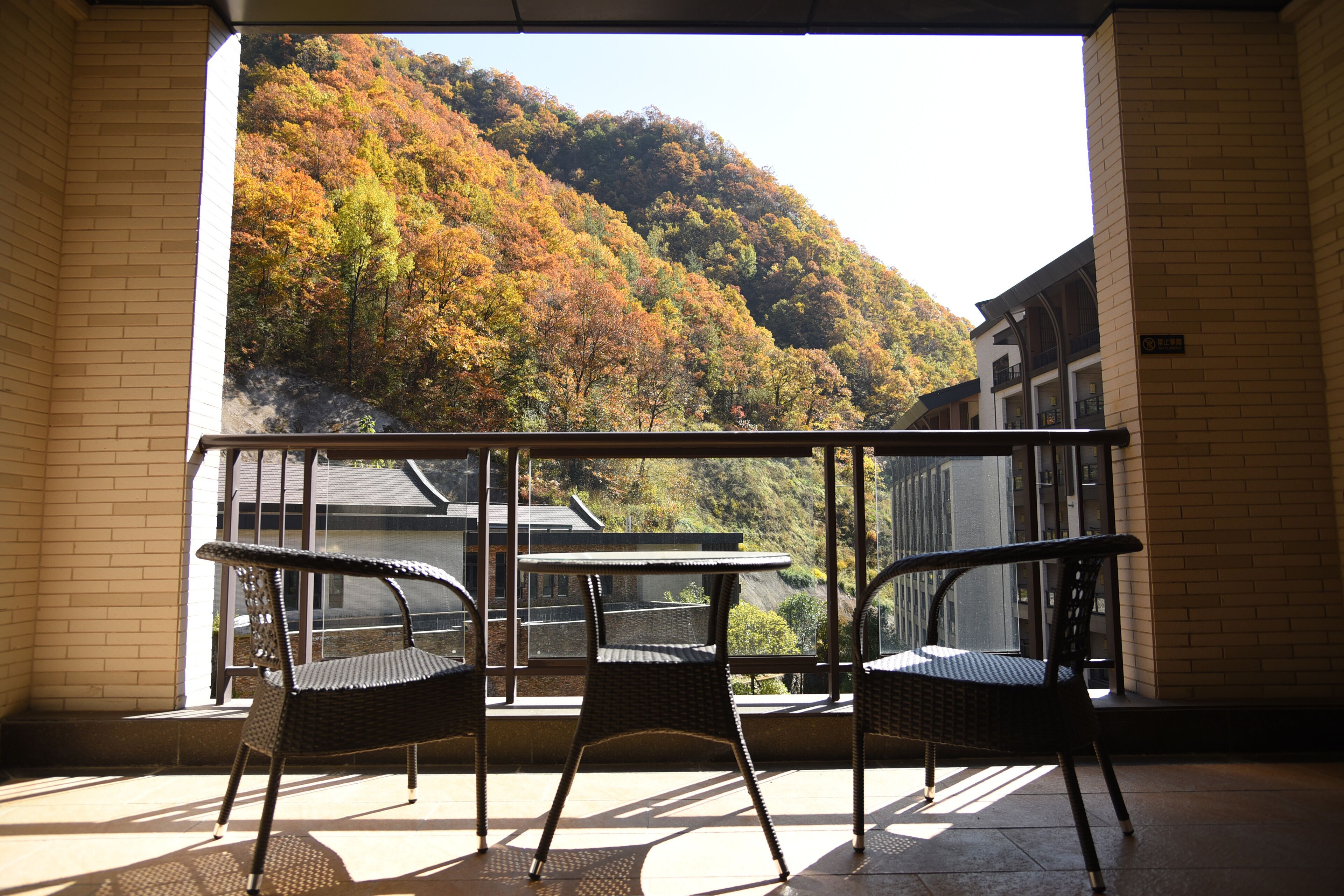 A balcony with two wicker chairs and a small table, overlooking a hillside with autumn foliage.