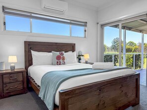 A bedroom with a large bed, wooden headboard, two bedside tables with lamps, a window, and a balcony with a view.