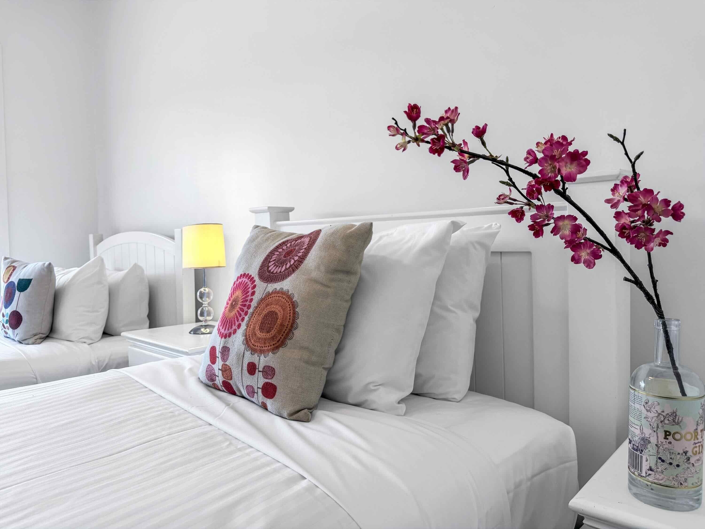 A hotel room with a bed, pillows, a bedside table with a lamp, and a vase with flowers.