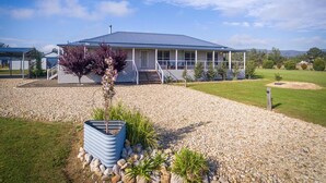 A single-story house with a covered porch, a gravel driveway, and a small garden with a tree and plants. - Kulang Lodge - A peaceful getaway on the outskirts of the village (Broke)