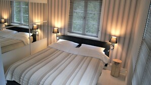 Hotellihuone, jossa on suuri sänky, kaksi yöpöytälamppua, peili ja ikkuna, jossa on sälekaihtimet.