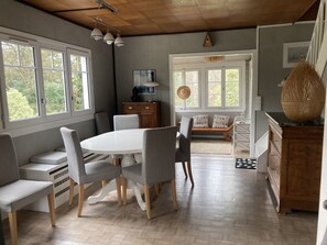Un espace repas comprenant une table ronde et des chaises, un banc en bois, et une vue sur l’extérieur grâce à de grandes fenêtres.
