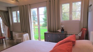Quarto com uma cama grande, penteadeira e uma cadeira. Há duas janelas com cortinas e vista para o verde lá fora.
