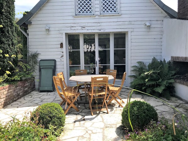 A white house with a patio featuring a wooden table and chairs, a brick wall, and a green door.