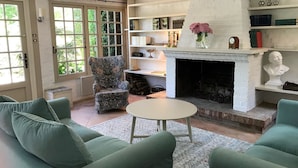 Una sala de estar acogedora con chimenea, sofás verdes, un sillón estampado y una estantería con libros y objetos decorativos.