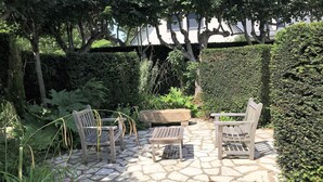 Un patio de piedra con muebles de madera, rodeado de exuberante vegetación y un seto.