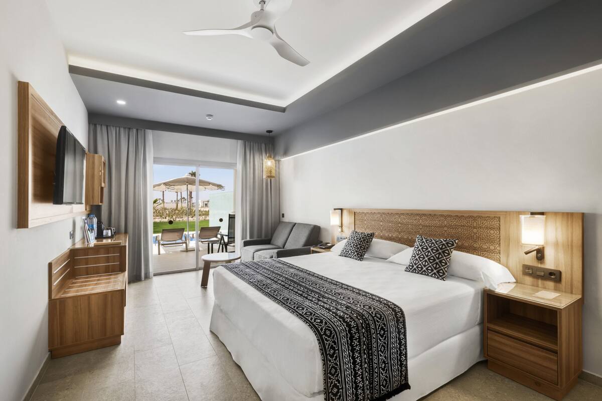 A modern hotel room with a large bed, a desk, a chair, a small table, and a view of the outdoors.