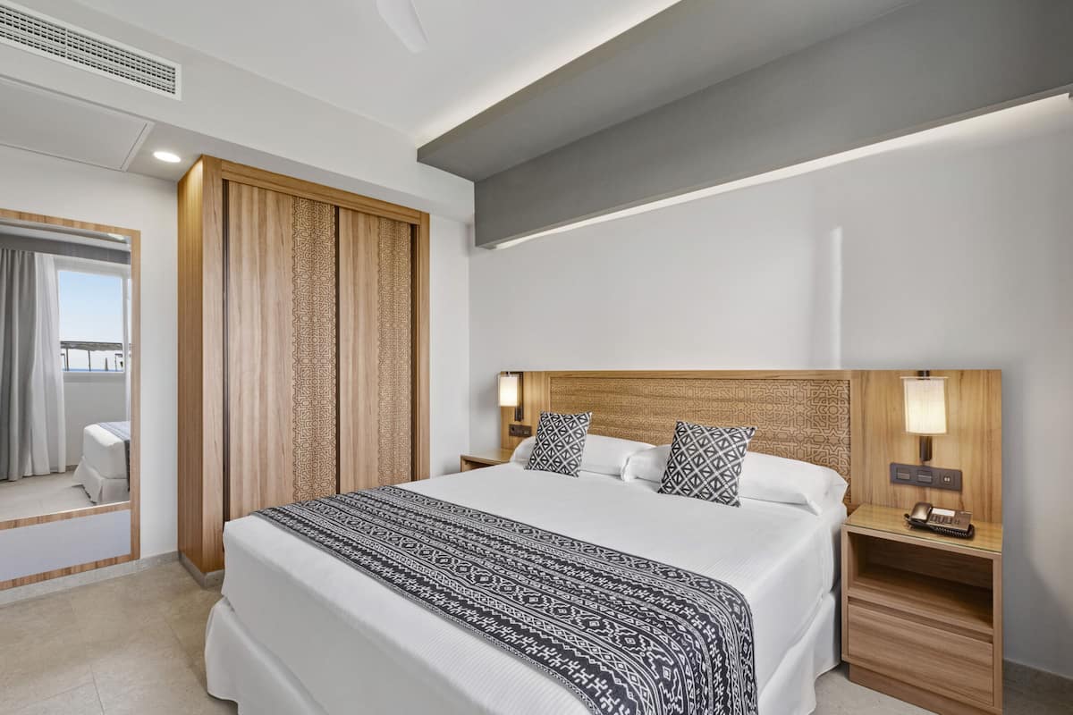 A modern hotel room with a large bed, wooden headboard, bedside lamps, and a small desk with a telephone.