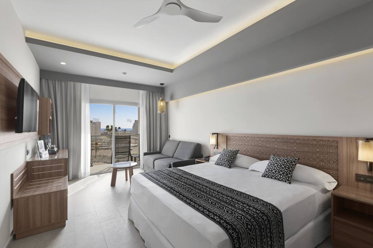 A modern hotel room with a large bed, a TV, a desk, and a balcony with a view.