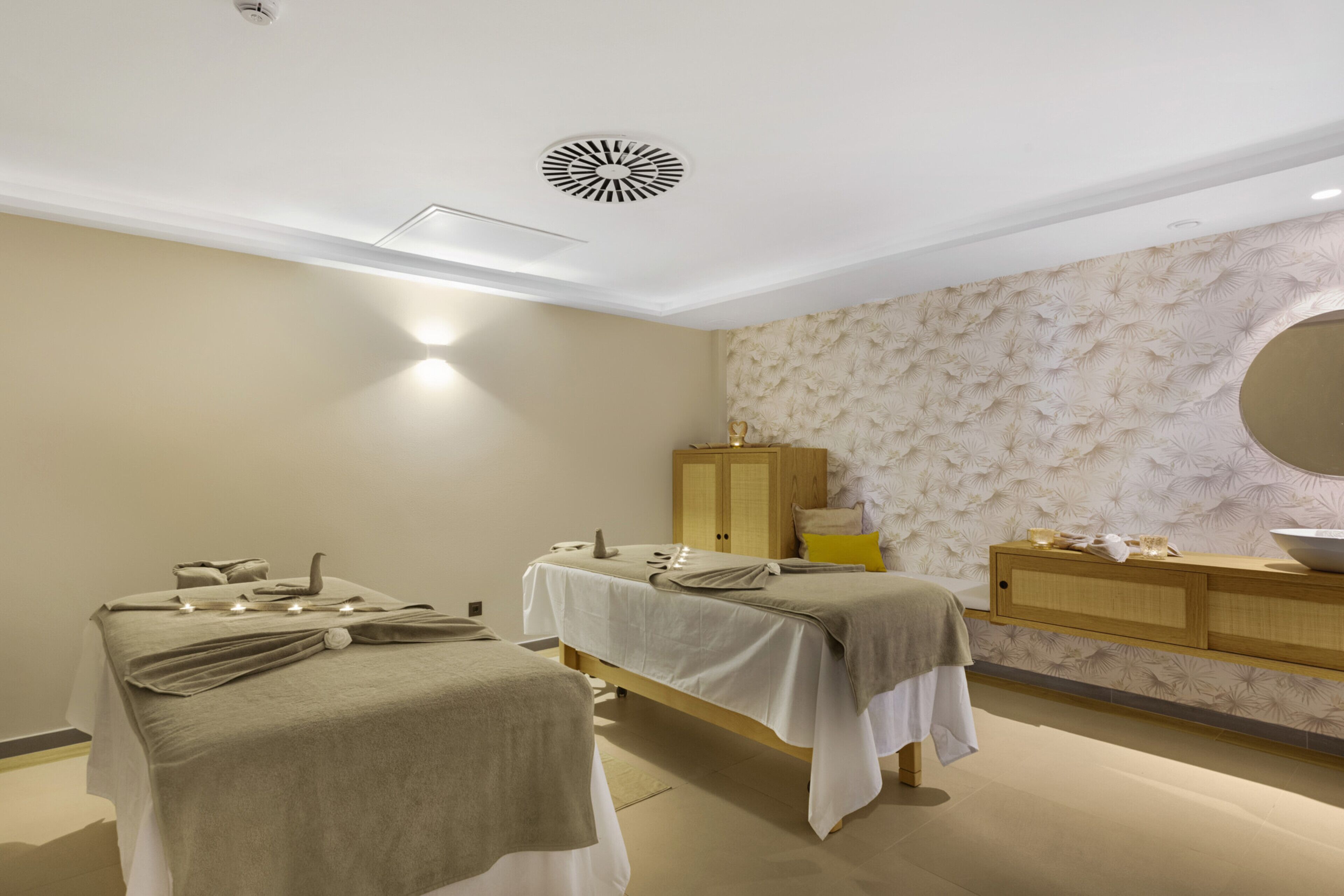 A spa room with two massage tables, a wooden cabinet, and a round mirror.