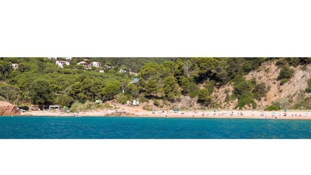 Una playa costera con casas en una colina, un mar azul cristalino y gente en la playa.