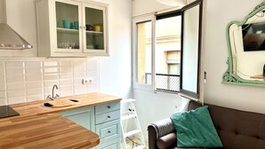 Cocina compacta con encimera de madera, un mueble blanco con puertas de cristal, una ventana con vistas a edificios y un sofá marrón con un cojín verde.