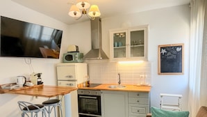 Una cocina compacta con encimera de madera, horno empotrado, microondas y lavabo. Hay taburetes de bar y un cojín verde sobre un sofá marrón.