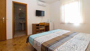 Quarto com cama, escrivaninha, cadeira, televisão e banheiro visível por uma porta aberta.