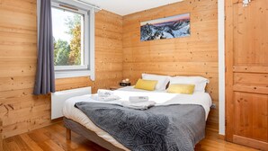 A wooden-paneled bedroom with a bed, pillows, a nightstand, a window, and a painting on the wall. - Le Paradis 25 Apartment -  an apartment that sleeps 6 guests in 3 bedrooms (Chamonix-Mont-Blanc)