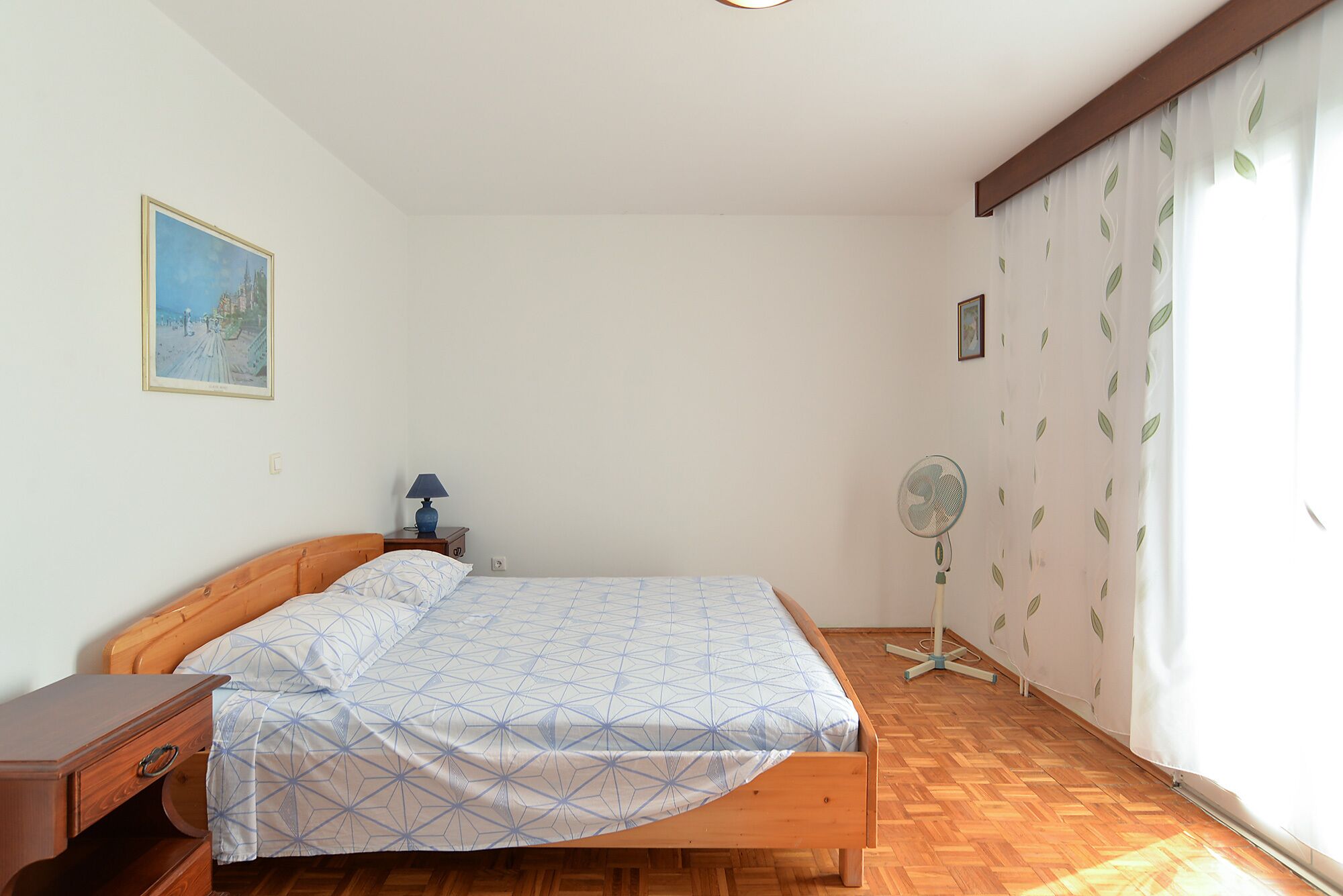 Een slaapkamer met een houten bed, nachtkastje, lamp, ventilator en een raam met gordijnen.
