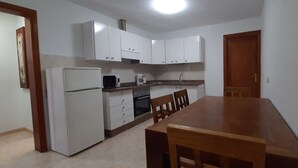Ett kompakt kök med vita skåp, ett kylskåp, en mikrovågsugn, en ugn och ett matbord med stolar.