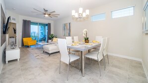 A modern dining area with a white table set for four, surrounded by white chairs. A ceiling fan and chandelier are visible above the table.