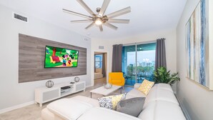 A modern living room with a flat-screen TV, white sofas, a yellow armchair, and a ceiling fan.