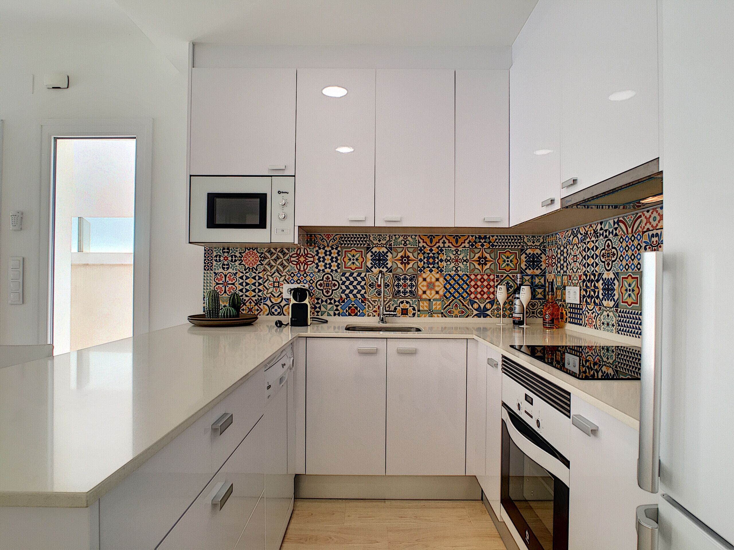Une cuisine moderne avec des meubles blancs, un four à micro-ondes et un dosseret de carrelage aux couleurs vives.